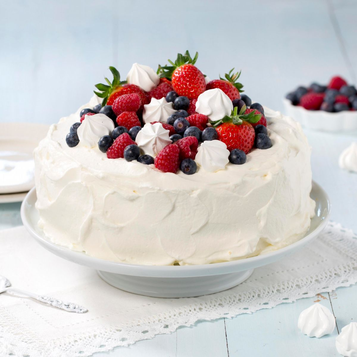 Bildet viser en kake dekket i hvit krem som står på et fat. Kaken er toppen med friske blåbær, bringebær og jordbær. I tillegg ligger det hvite pikekyss/marengs mellom den fargerike frukten på toppen.