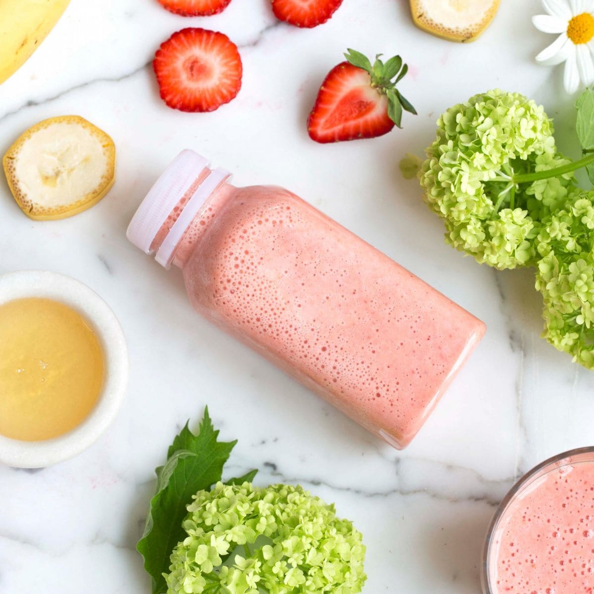 Bildet viser en flaske liggende på et bord. Innholdet i flasken er lyserosa og inneholder smoothie. Rundt flasken ligger det ferske bær som jordbær og ingredienser som banan.