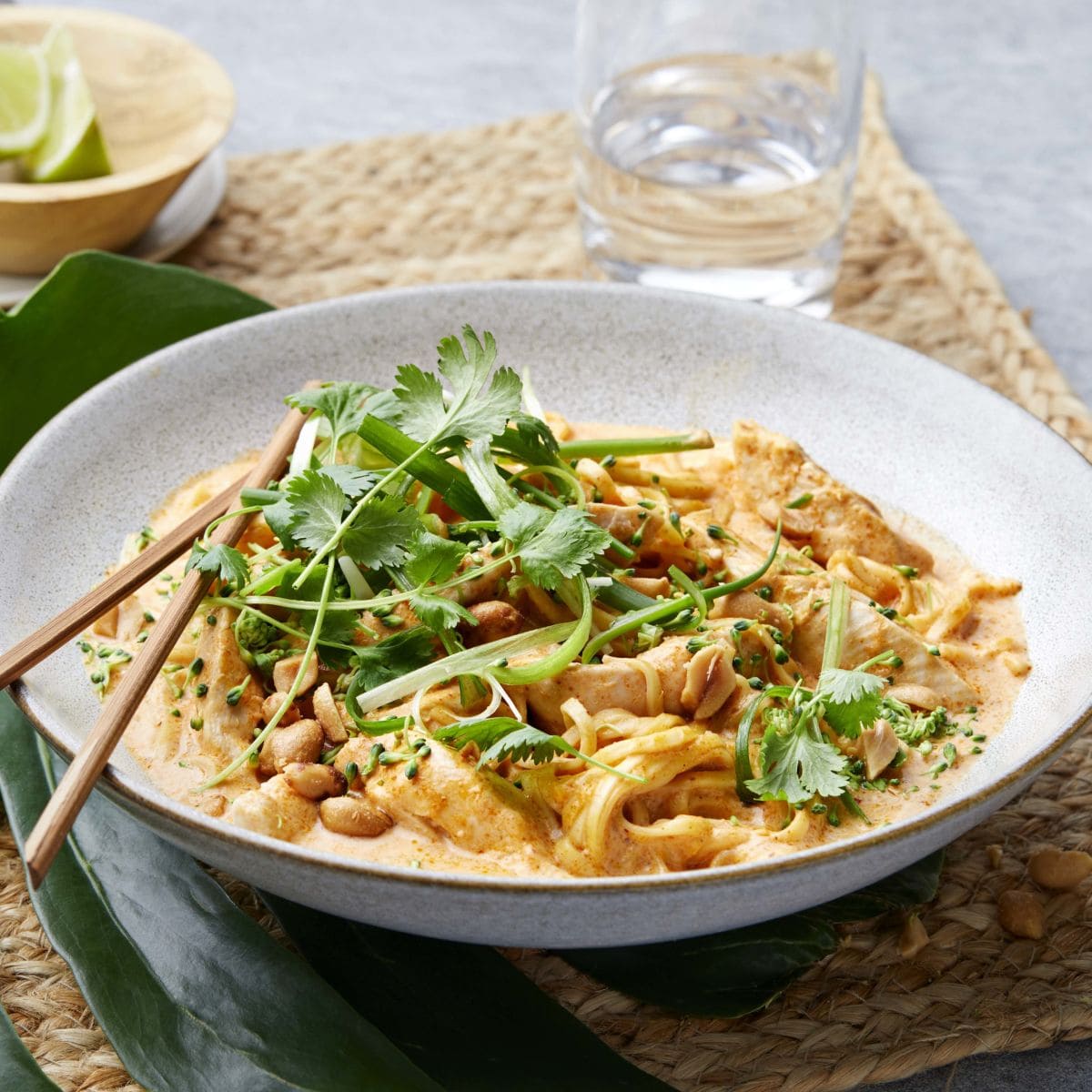 Et fargerikt bilde av en asiatisk red currygryte med kylling, servert i en dyp tallerken. Gryten er fylt med saftige kyllingstrimler, eggnudler, og en rik rød currysaus laget med kokosmelk og crème fraîche. Retten er toppet med finstrimlet vårløk, raspet brokkoli, grovhakkede peanøtter og fersk koriander, som gir en frisk og sprø tekstur. I bakgrunnen kan man se ingredienser som lime, chili og sitrongress, som fremhever de asiatiske smakene i retten.