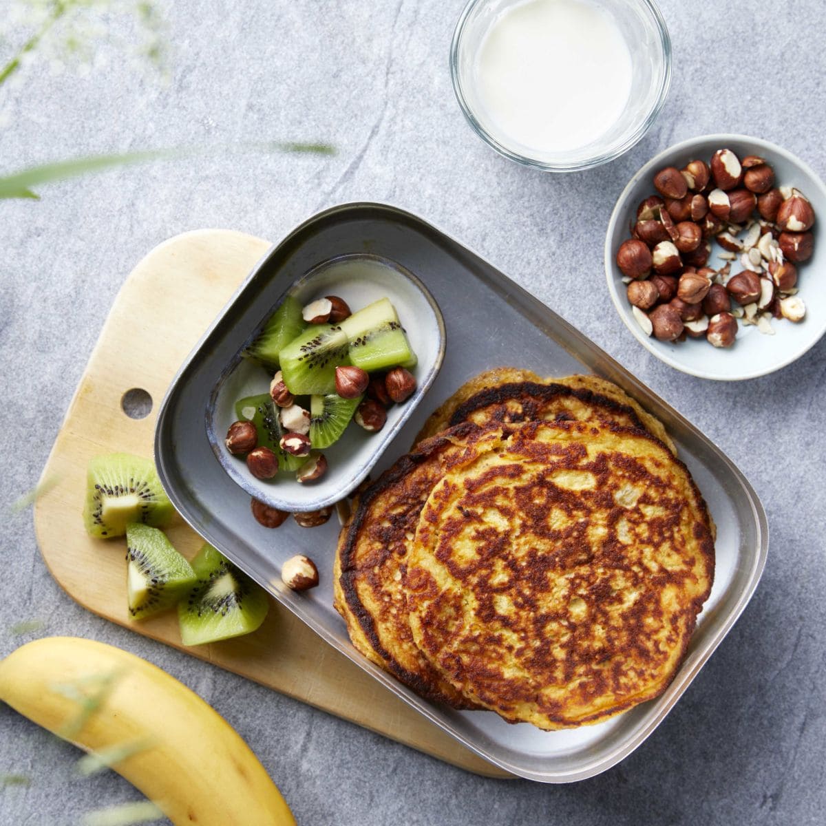Bildet viser en matboks på en grå kjøkkenbenk. Oppi matboksen ligger det to runde pannekaker og en liten skål med oppkuttet grønn kiwi og nøtter. Ved siden av matboksen ligger det en gul banan, flere kiwi biter, en skål med nøtter og en iskaldt glass melk.