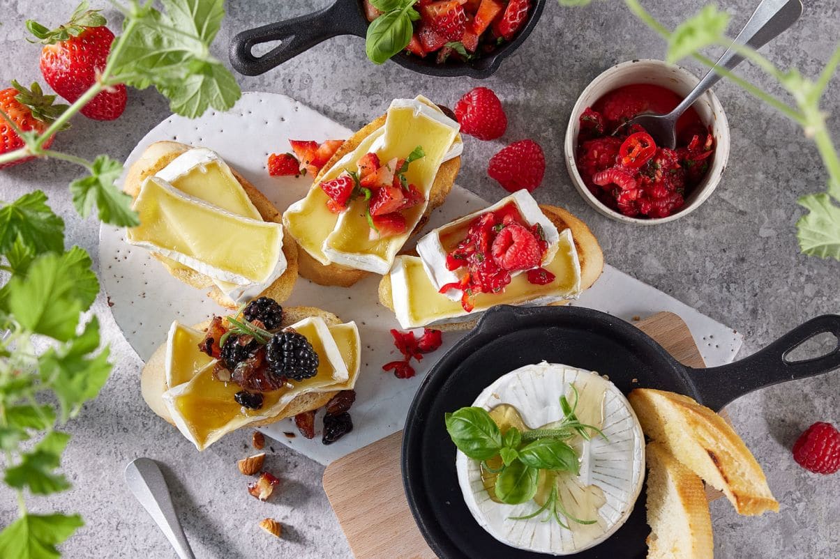 Bildet viser fristende små biter av lyst brød toppet med bakt brie som er til dels smeltet. På toppen av de ulike bitene ligger det friske bær som røde jordbær og sorte bjørnebær. Nederst i venstre hjørne kan man se en hel brie i en sort liten stekepanne.