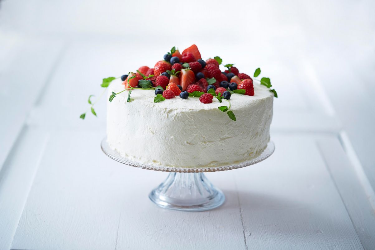 Bildet viser en hvit kake dekket i krem som står på et stetfat i krystall. Kremkaken er toppet med friske bær som blåbær, jordbær og bringebær. Helt på topp et det pyntet med noen friske, grønne mynteblader.