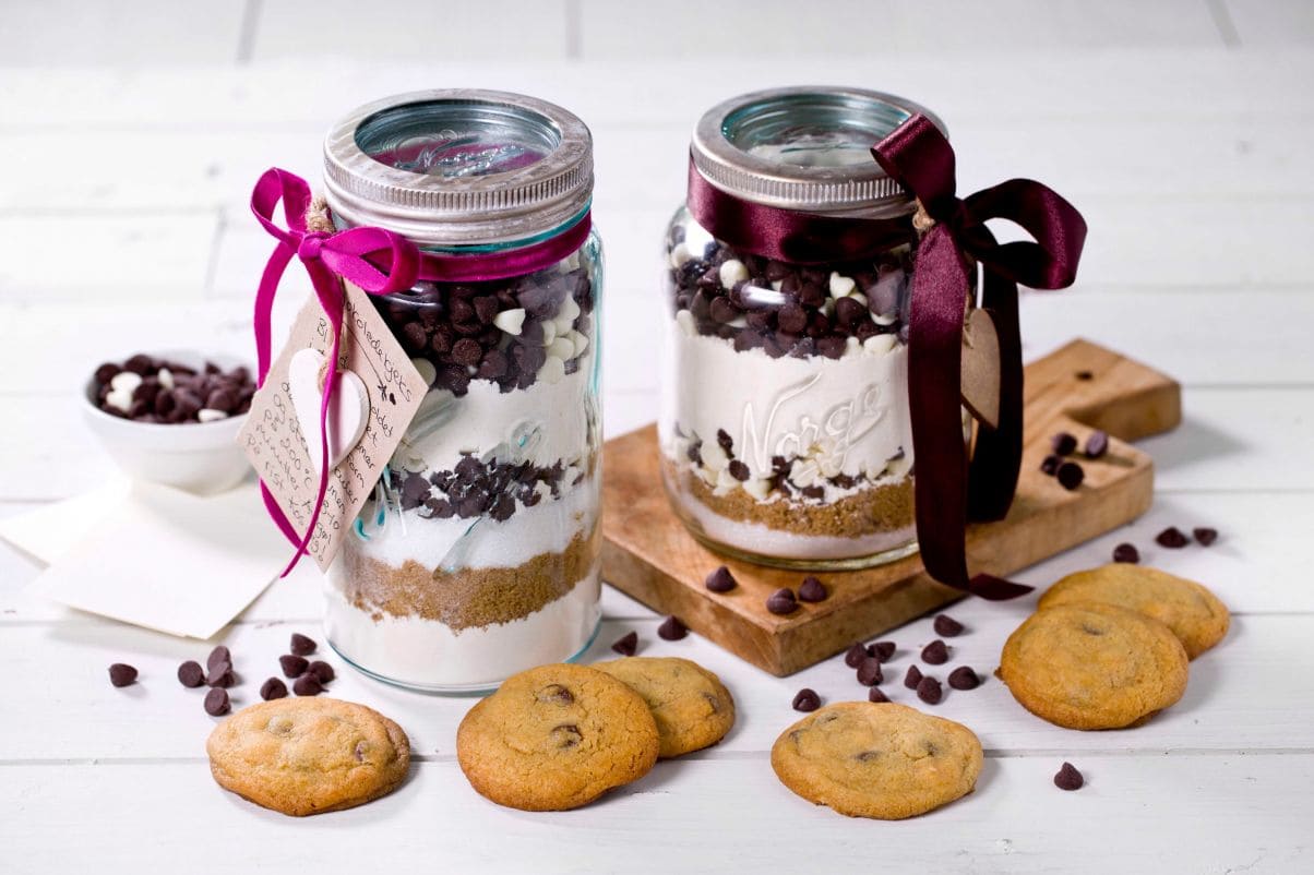Bildet viser en glasskrukke cookies stå på en benk. Foran glasskrukken står det en cookie opp på høykant støttet inntil et glass melk. I bakgrunn skimtes et kjøkken.