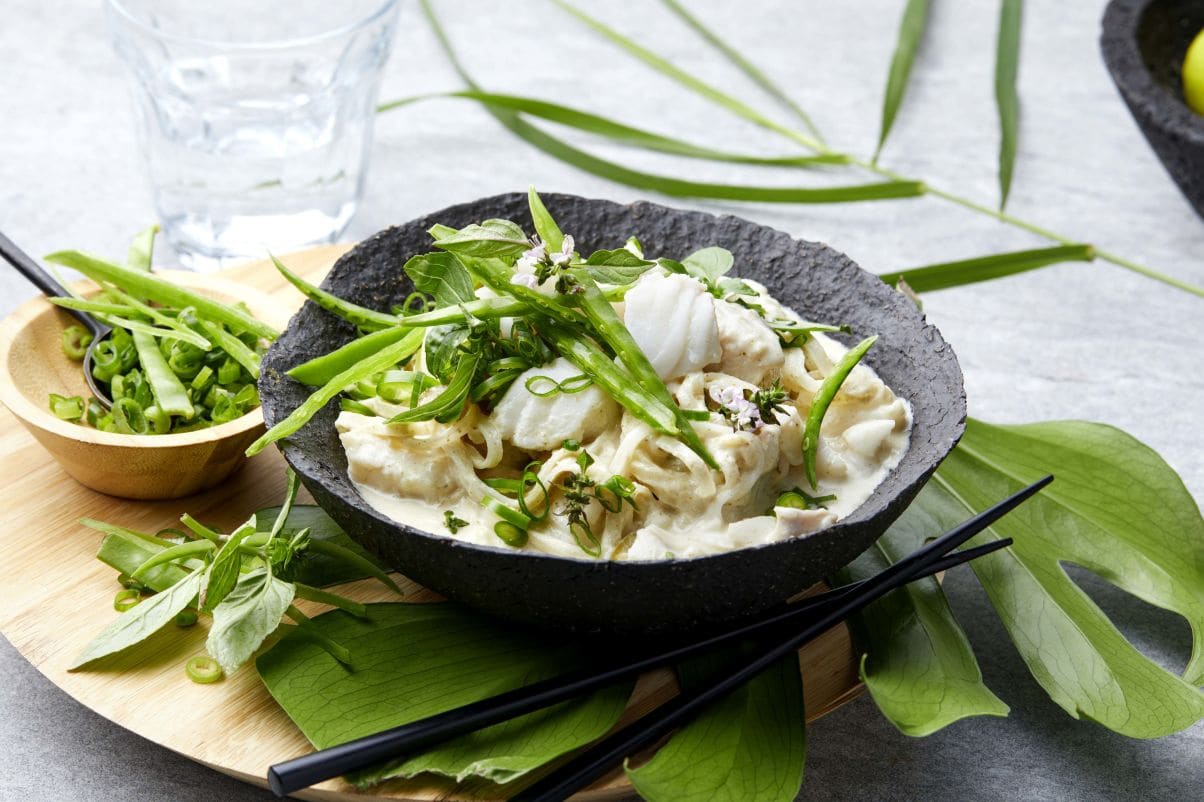 Bildet viser en svart bolle med en kremet curryrett, toppet med grønne urter og vårløk. Bollen står på et rundt trebrett sammen med en liten skål fylt med oppskårne grønnsaker. Ved siden av ligger et par svarte spisepinner og noen grønne blader som dekorasjon. I bakgrunnen er det et glass vann og en tallerken med limebåter.