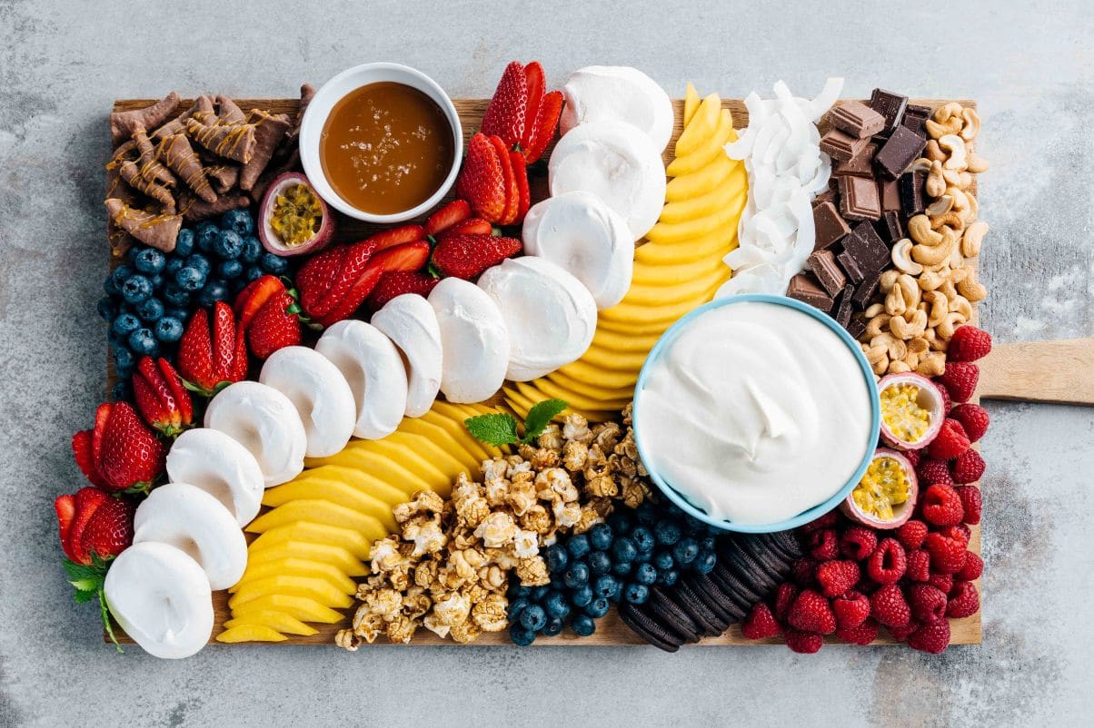 Bildet viser et stort brett med flere små pavlovaer, også kjent som pavlova-tapas eller pavlova board. Hver minipavlova består av en sprø marengsbunn med en liten grop i midten, fylt med krem og toppet med ulike frukter og bær. På brettet er det en rekke forskjellige toppinger, inkludert skiver av jordbær, mango, pasjonsfrukt, blåbær, bringebær, og karamellisert popcorn. Det er også biter av melkesjokolade og mørk sjokolade, samt knuste oreokjeks og Smash®. I midten av brettet er det en skål med pisket krem blandet med vaniljekrem. Fargene i bildet er lyse og fristende, med en kombinasjon av hvit marengs, fargerike frukter og bær, og sjokolade.