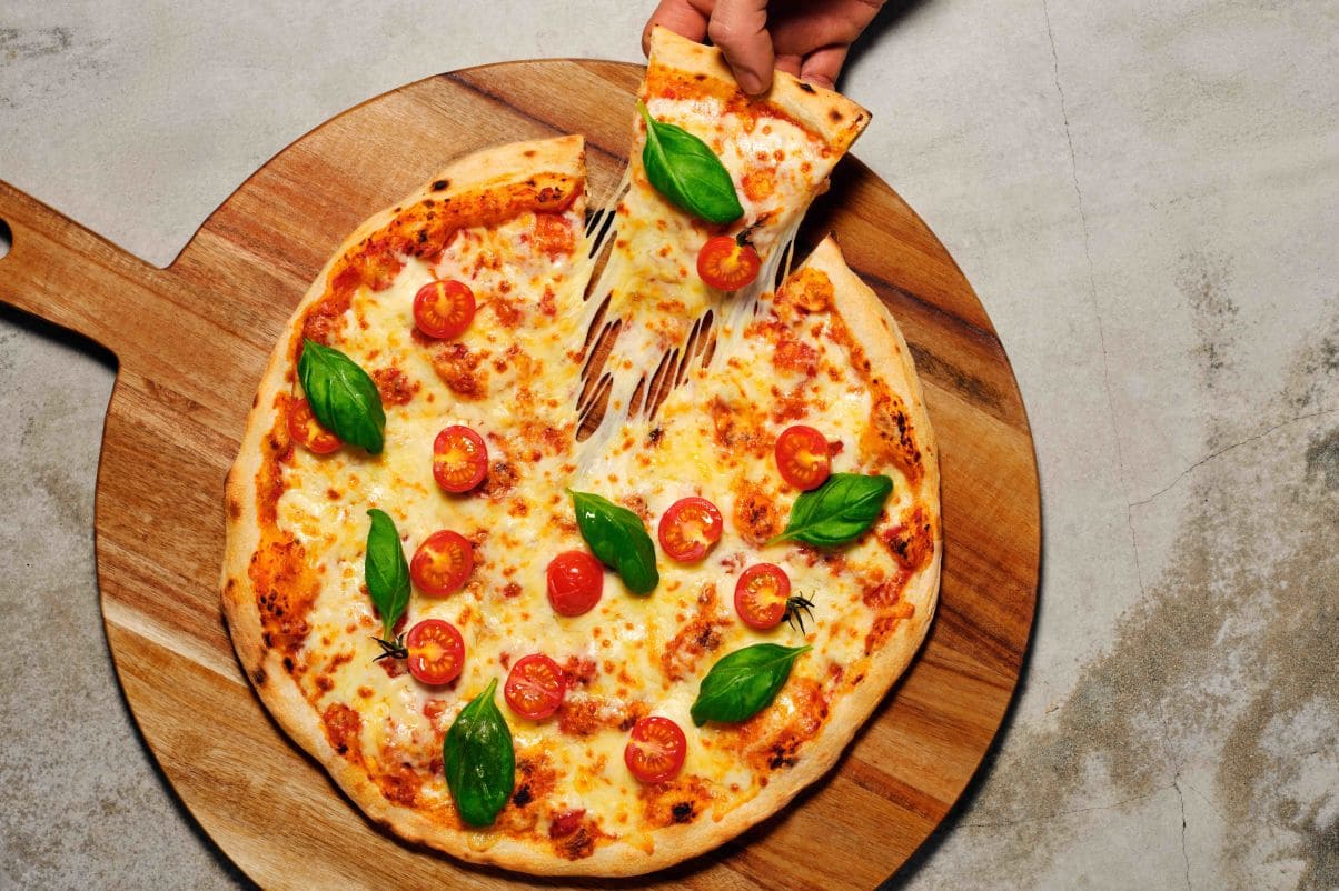 Pizza Margherita toppet med delte cherrytomater og basilikumblader på en fjøl. En hånd kommer inn i bildet og tar et pizzastykke. 