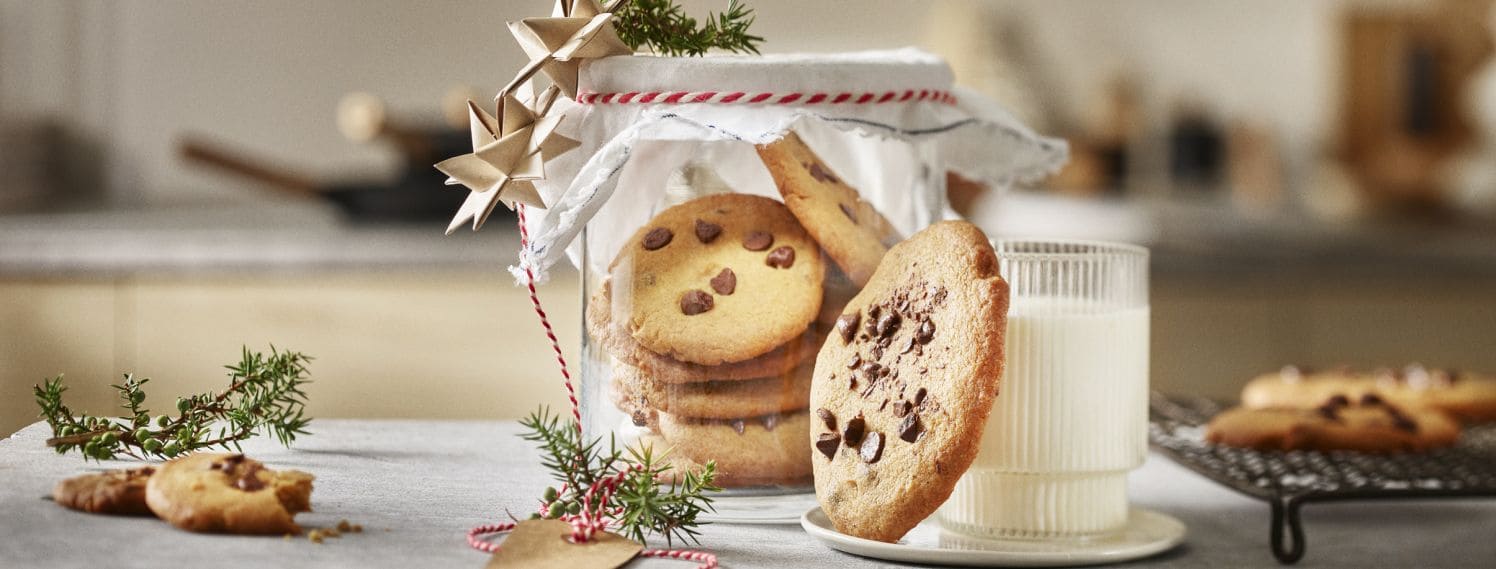 Glasskrukke med sjokoladecookies. Krukken er pyntet med litt granbar og julestjerner av papir. Foran står et glass melk med en sjokoladecookie. 