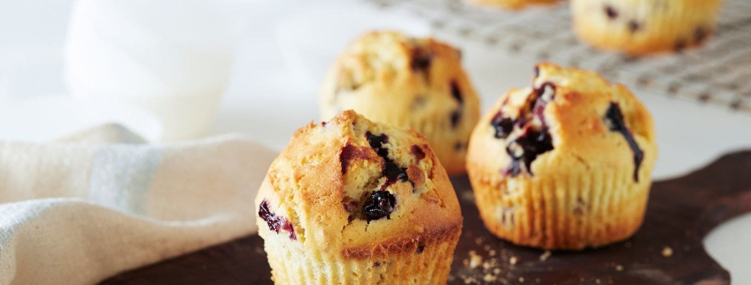 Bildet viser en tallerken med flere blåbærmuffins, som er plassert på en lys bakgrunn. Muffinsene er gyllenbrune på toppen og har synlige blåbær som gir dem en fristende, saftig utseende. Hver muffin er pent formet og ser ut til å være myke og luftige.