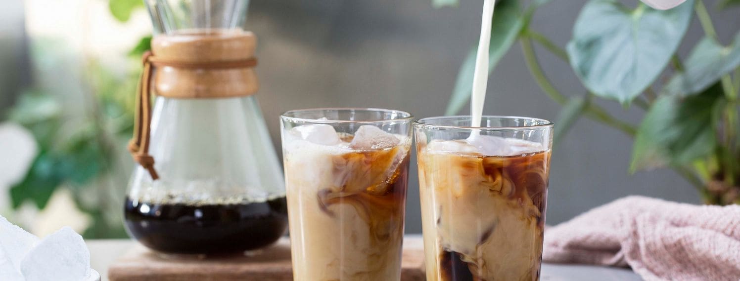 Bildet viser to glass som står på en kjøkkenbenk. Glassene er fulle og inneholder kaffe, isbiter og melk. Kombinasjonen gjør at det er mønster i glasset. I det ene glasset helles det hvit melk fra toppen av bildet. Dette skaper et pent mønster i glasset, blant den mørke kaffen og isbitene.