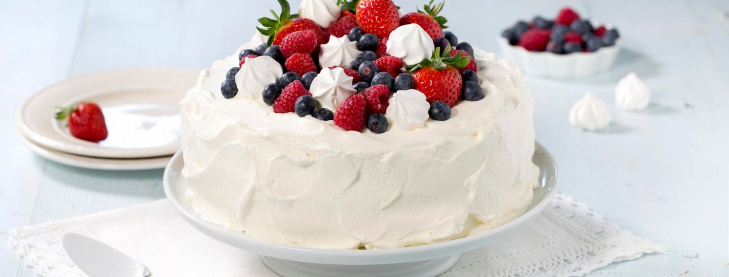 Bildet viser en kake dekket i hvit krem som står på et fat. Kaken er toppen med friske blåbær, bringebær og jordbær. I tillegg ligger det hvite pikekyss/marengs mellom den fargerike frukten på toppen.