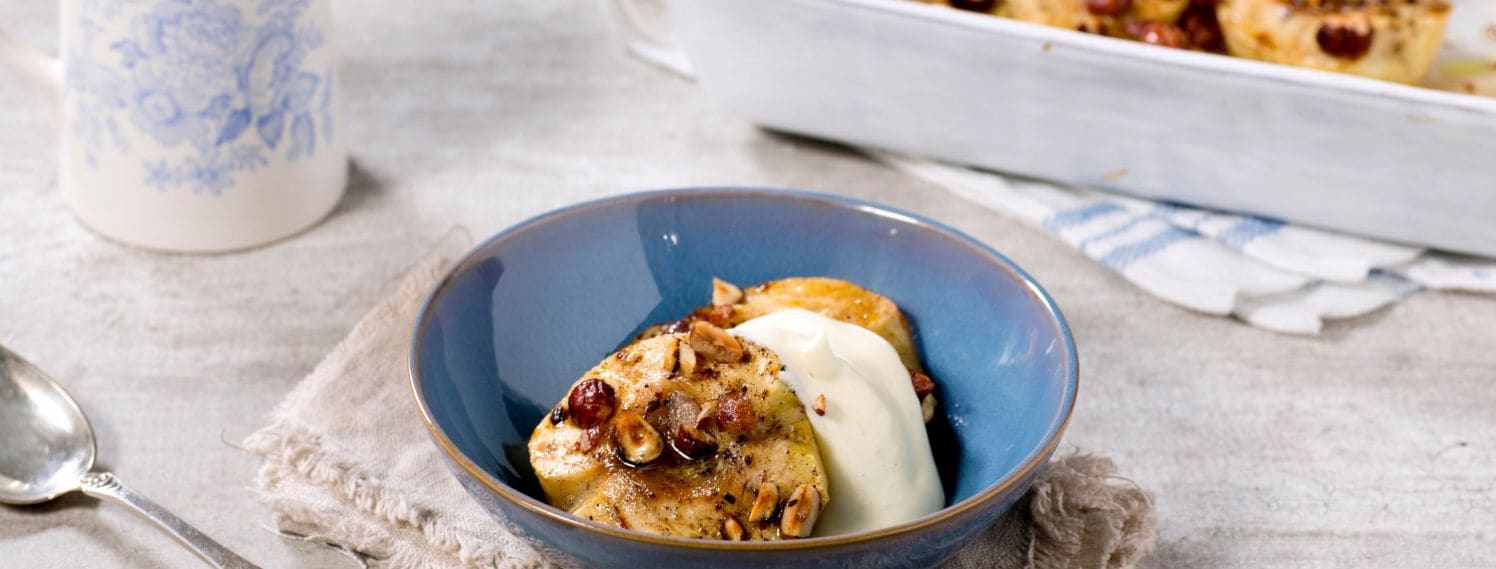 Bildet viser en dessert bestående av bakte epler servert med luftig vaniljesaus. Eplene er delt i to og bakt til de er myke, og de er plassert i en hvit, dyp tallerken. Hver eplehalvdel er toppet med en gyllenbrun blanding av kanel, brunt sukker og hakkede nøtter, som gir en sprø tekstur og en rik smak. Rundt eplene er det en generøs mengde luftig vaniljesaus, som er pisket til en lett og kremet konsistens. Sausen har en lys gul farge som står i kontrast til de karamelliserte eplene.