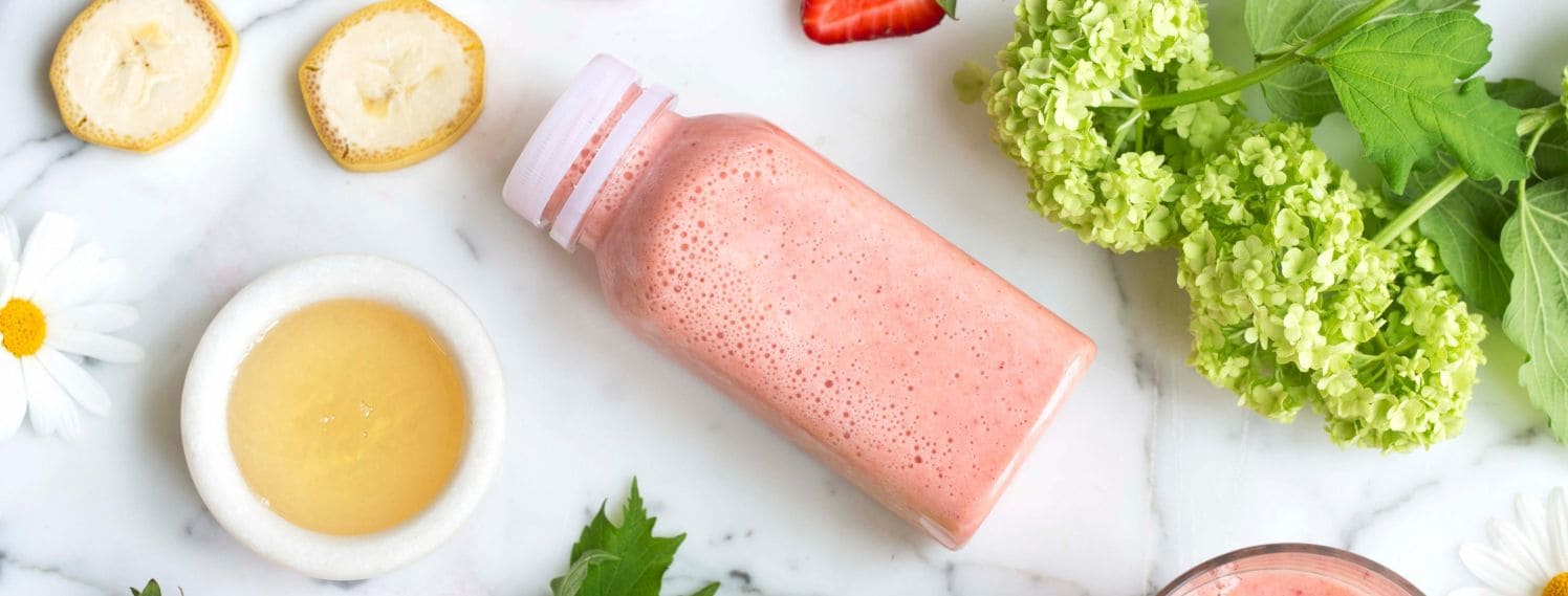 Bildet viser en flaske liggende på et bord. Innholdet i flasken er lyserosa og inneholder smoothie. Rundt flasken ligger det ferske bær som jordbær og ingredienser som banan.