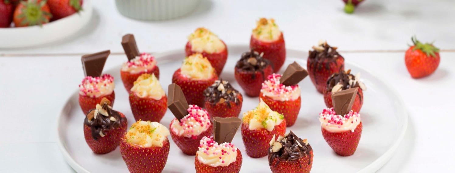 Bildet viser en delikat dessert bestående av fylte jordbær. Jordbærene er delt i to og fylt med en kremet blanding av kremost og melis. Hvert jordbær er toppet med en liten mynteblad, som gir en frisk kontrast til den søte fyllet. Jordbærene er pent arrangert på en hvit tallerken, og i bakgrunnen kan man se en lys og nøytral overflate som fremhever de røde bærene. Dette bildet gir en følelse av friskhet og sommer, og viser en enkel, men elegant dessert som er perfekt for enhver anledning.