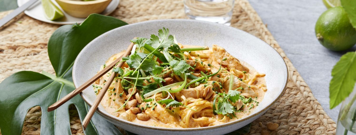 Et fargerikt bilde av en asiatisk red currygryte med kylling, servert i en dyp tallerken. Gryten er fylt med saftige kyllingstrimler, eggnudler, og en rik rød currysaus laget med kokosmelk og crème fraîche. Retten er toppet med finstrimlet vårløk, raspet brokkoli, grovhakkede peanøtter og fersk koriander, som gir en frisk og sprø tekstur. I bakgrunnen kan man se ingredienser som lime, chili og sitrongress, som fremhever de asiatiske smakene i retten.