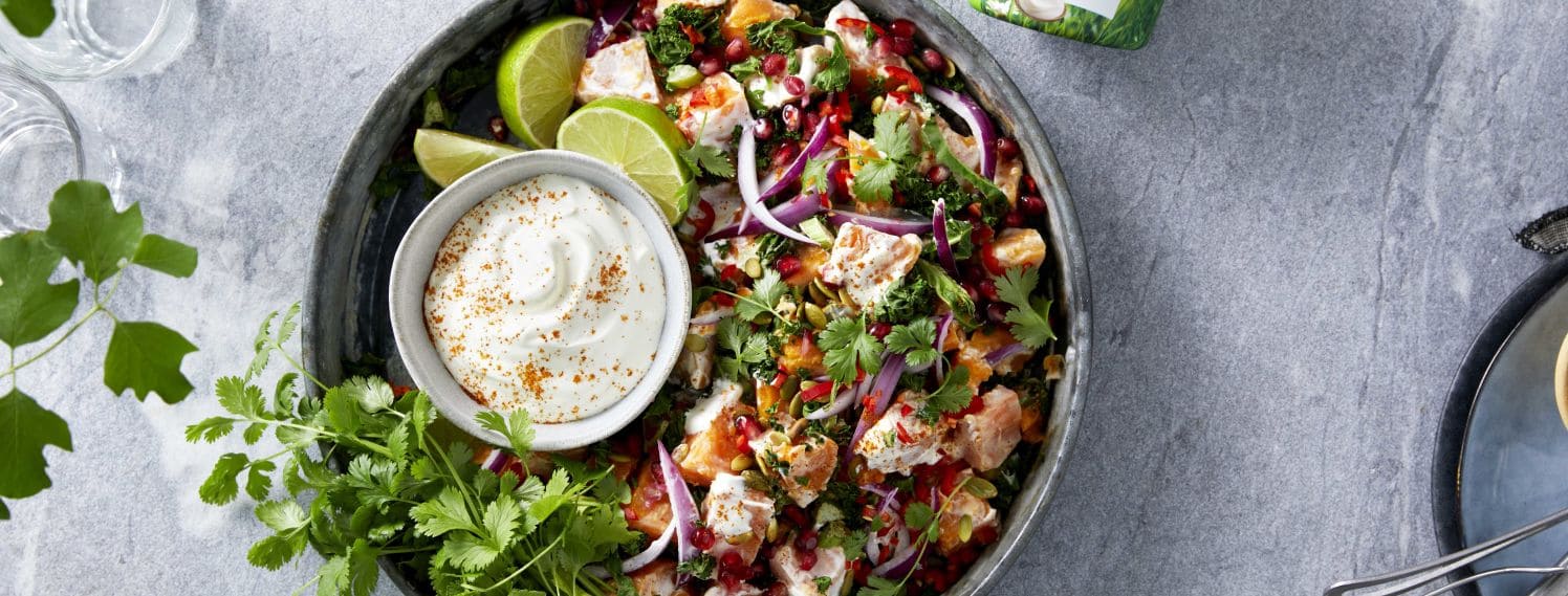 Bildet viser en rund skål fylt med kremet søtpotetsalat. I salaten ser vi også limebåter, en skål med rømmedressing, frisk persille i grønt, lilla rødløk kuttet i tynne skiver og røde granateplekjerner.