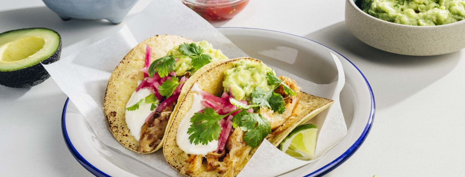 Bildet viser en tallerken med to kyllingtacos. De er toppet med lillafarget syltet rødløk og grønn avokoado.