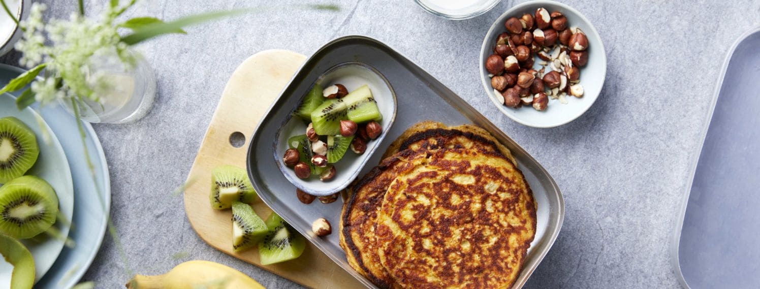 Bildet viser en matboks på en grå kjøkkenbenk. Oppi matboksen ligger det to runde pannekaker og en liten skål med oppkuttet grønn kiwi og nøtter. Ved siden av matboksen ligger det en gul banan, flere kiwi biter, en skål med nøtter og en iskaldt glass melk.