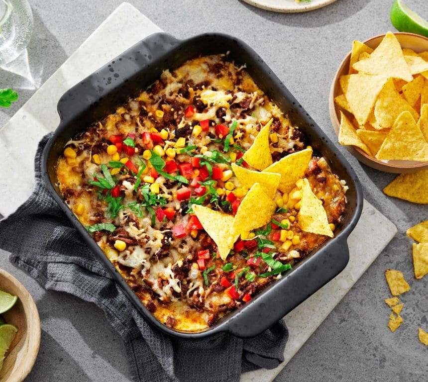 En svart ildfast form fylt med tacogryte: kjøttdeig og ost, toppet med mais, paprika, hakket koriander og nachochips. Ved siden av står en skål med flere nachochips og en skål med limebåter