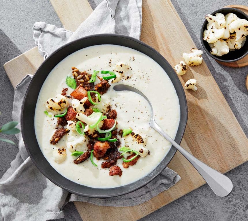 Bildet viser en rund, mørk skål fylt med hvit blomkålsuppe med sprø, brun bacon og grønn purre. Ved siden av skålen ligger en spiseskje.