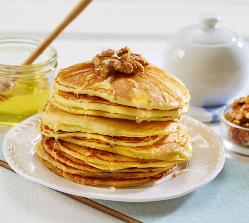 Bildet viser en stabel med tykke, amerikanske pannekaker. Pannekakene er toppet med lys lønnesirup og brune valnøtter.