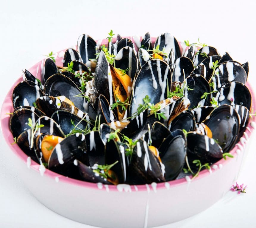 En tallerken med nydampede blåskjell, servert med en skål crème fraîche- og hvitløksdipp. Bakgrunnen viser en lys bordflate.