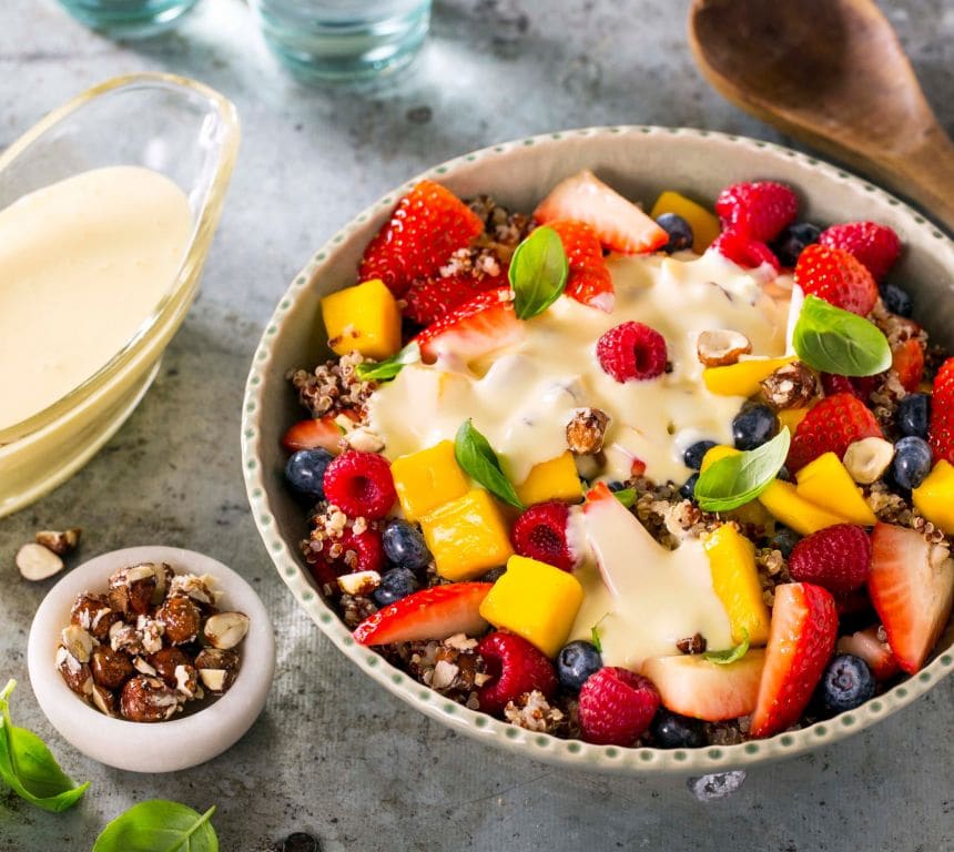 En bolle med fruktsalat med jordbær, bringebær, blåbær og mango, toppet med basilikum, vaniljesaus og ristede hasselnøtter