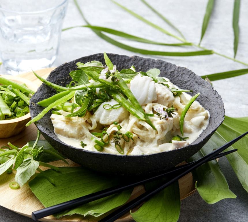 Bildet viser en svart bolle med en kremet curryrett, toppet med grønne urter og vårløk. Bollen står på et rundt trebrett sammen med en liten skål fylt med oppskårne grønnsaker. Ved siden av ligger et par svarte spisepinner og noen grønne blader som dekorasjon. I bakgrunnen er det et glass vann og en tallerken med limebåter.