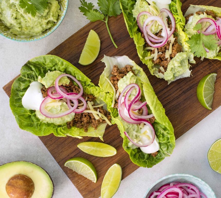 Bildet viser tre hjertesalat blader toppet med krydret tacokjøttdeig, guacamole og revet ost. Ved siden av salatbladene med fyll ligger det en halv grønn avokado.