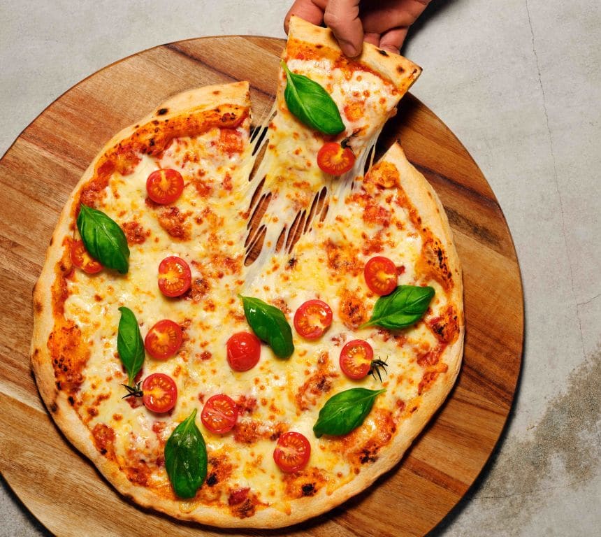 Pizza Margherita toppet med delte cherrytomater og basilikumblader på en fjøl. En hånd kommer inn i bildet og tar et pizzastykke. 