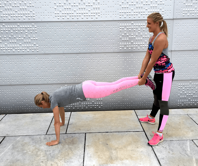 To jenter trener sammen. Den ene gjør knebøy og holder bena til den andre. Den andre tar pushups.