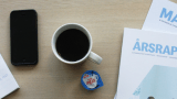 Blokk, kulepenn, mobiltelefon, kaffekopp, TINE Kaffefløte, TINEs årsrapport og TINE Magasinet liggende på bord